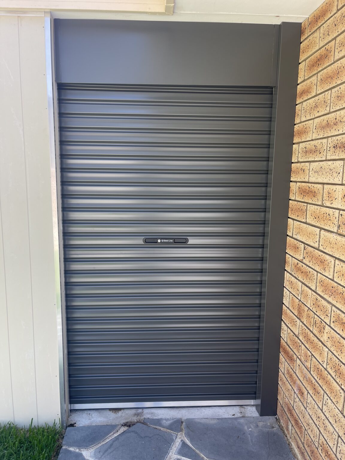 Grey roller door fitted to a narrow garage opening beside a brick wall