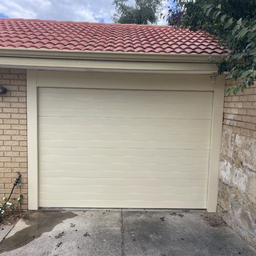 Cream sectional garage door on a single garage with a red tile roof and brick wall